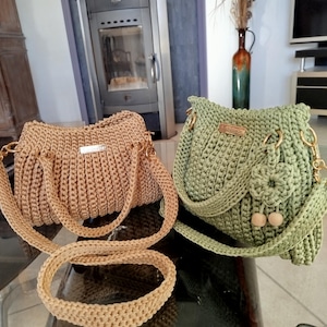 Puede incluir: Dos bolsos de crochet hechos a mano. Uno es de color beige con una correa larga y asas cortas, el otro es verde con una solapa, cuentas de madera y una correa de cadena dorada. Ambos bolsos tienen herrajes dorados.