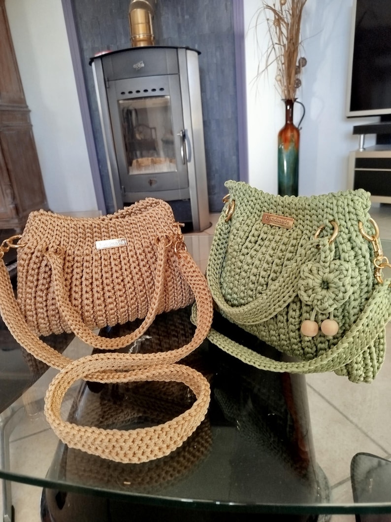 Puede incluir: Dos bolsos de crochet hechos a mano. Uno es de color beige con una correa larga y asas cortas, el otro es verde con una solapa, cuentas de madera y una correa de cadena dorada. Ambos bolsos tienen herrajes dorados.