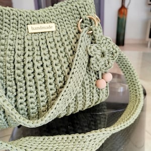 Puede incluir: Un bolso de mano de ganchillo hecho a mano, verde salvia, con forma rectangular. El bolso presenta una correa larga trenzada y herrajes dorados. Una peque&ntilde;a etiqueta de madera con la palabra "handmade" est&aacute; adherida al frente. La correa est&aacute; adornada con una flor y dos peque&ntilde;as cuentas rosas.