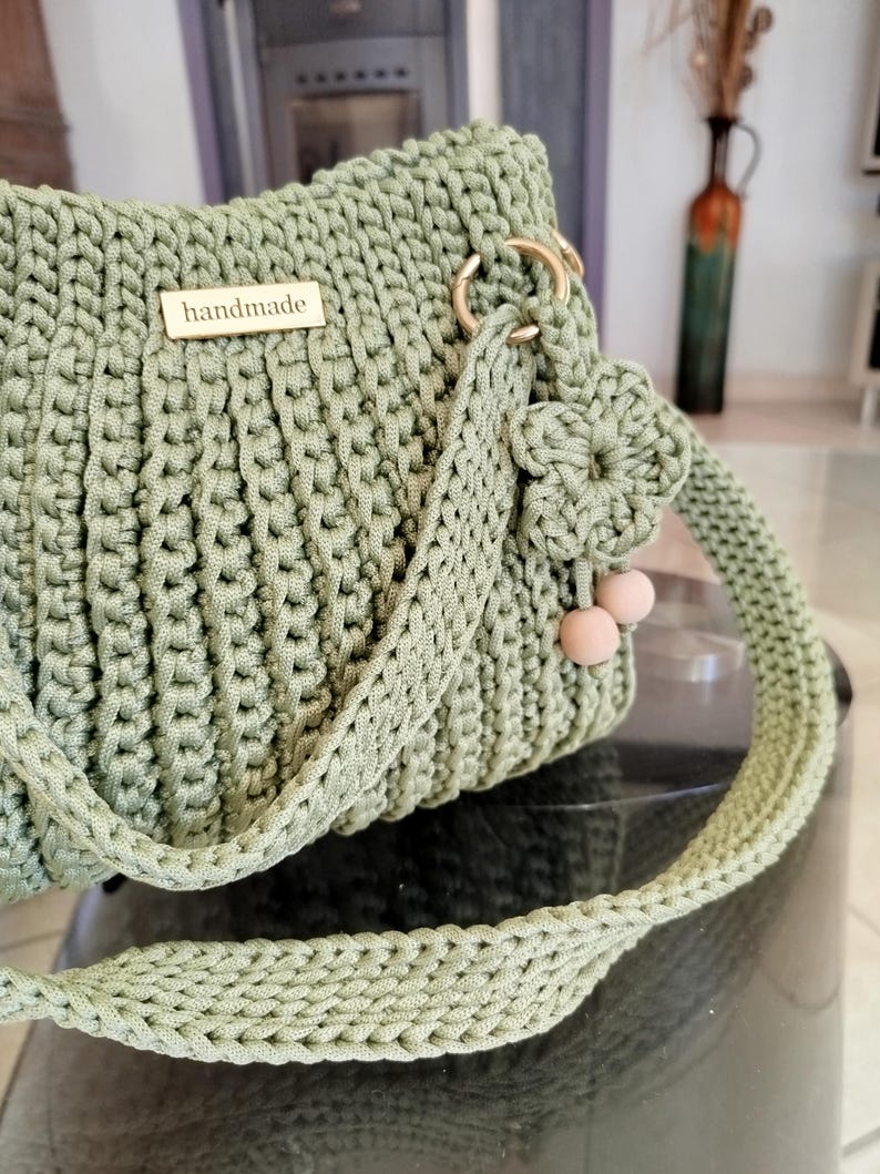 Puede incluir: Un bolso de mano de ganchillo hecho a mano, verde salvia, con forma rectangular. El bolso presenta una correa larga trenzada y herrajes dorados. Una peque&ntilde;a etiqueta de madera con la palabra "handmade" est&aacute; adherida al frente. La correa est&aacute; adornada con una flor y dos peque&ntilde;as cuentas rosas.