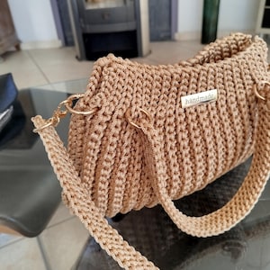 Puede incluir: Un bolso de mano de ganchillo color beige con herrajes dorados. El bolso presenta una textura tejida y una correa para el hombro. Una peque&ntilde;a etiqueta met&aacute;lica con la inscripci&oacute;n "handmade" est&aacute; adherida al frente. El bolso est&aacute; sobre una mesa de cristal.