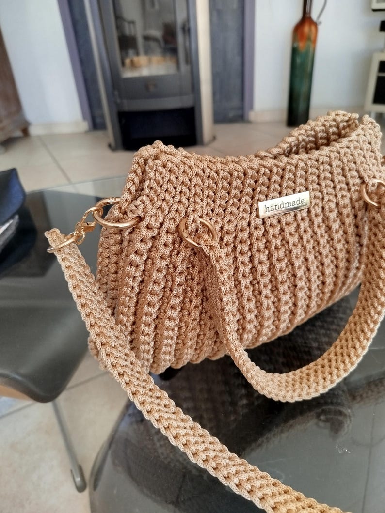Puede incluir: Un bolso de mano de ganchillo color beige con herrajes dorados. El bolso presenta una textura tejida y una correa para el hombro. Una peque&ntilde;a etiqueta met&aacute;lica con la inscripci&oacute;n "handmade" est&aacute; adherida al frente. El bolso est&aacute; sobre una mesa de cristal.