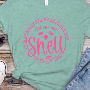May include: A light blue t-shirt with a pink circle design. The circle has the text "Just one more shell" and the names of different types of shells around the circle.