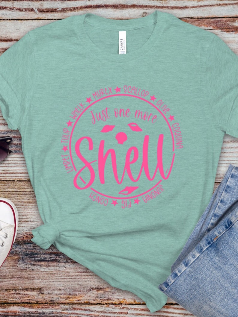 May include: A light blue t-shirt with a pink circle design. The circle has the text "Just one more shell" and the names of different types of shells around the circle.