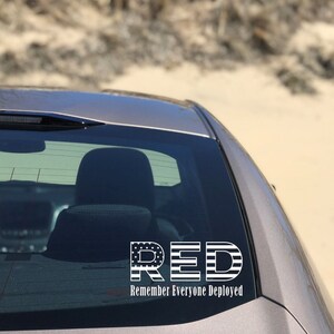 May include: A white vinyl car sticker with the acronym "RED" in large letters, with each letter designed to look like the American flag. Below the letters is the text "Remember Everyone Deployed".