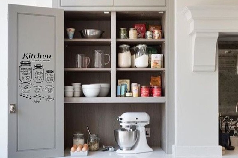 May include: A white kitchen cabinet with glass shelves filled with various food items and kitchen tools. The cabinet door has a black and white kitchen conversion chart sticker with the text "Kitchen Conversions".