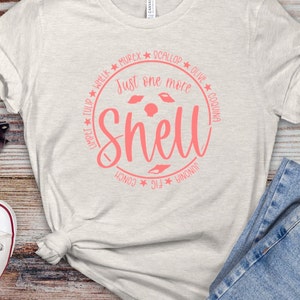 May include: A light gray t-shirt with a pink circle graphic that says "Just one more shell" with a variety of seashells around the text.