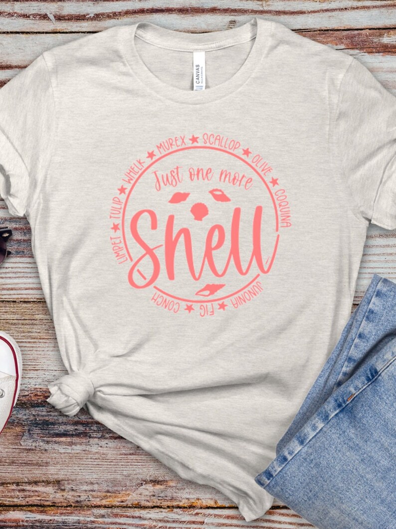 May include: A light gray t-shirt with a pink circle graphic that says "Just one more shell" with a variety of seashells around the text.