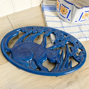 Puede incluir: Un salvamanteles ovalado de hierro fundido en color azul intenso con un diseño en relieve de un gato sentado entre flores y follaje. El salvamanteles tiene un acabado brillante y un patrón calado que muestra la figura felina de perfil.