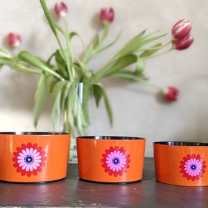 Op de afbeelding: Drie oranje decoratieve kommen met een retro bloemenontwerp. Elke kom heeft een grote bloem met roze en blauwe bloemblaadjes. De kommen zijn in een rij gerangschikt, met een vaas met tulpen op de achtergrond.