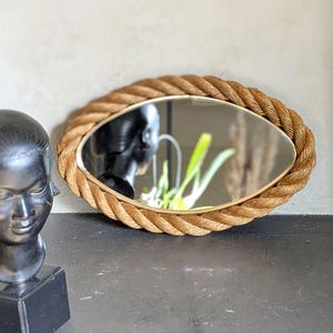 May include: Oval mirror with a rope-style frame. The mirror's frame is a light brown color, and the mirror itself reflects the surrounding environment. A black sculpture is visible to the left of the mirror.