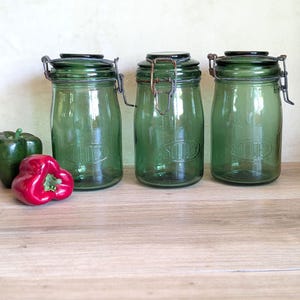 Peut inclure: Trois bocaux en verre vert avec couvercles à charnière et fermoirs métalliques. Les bocaux sont cylindriques avec une base légèrement arrondie. Un poivron rouge et un poivron vert sont à côté des bocaux. Sur une surface en bois.