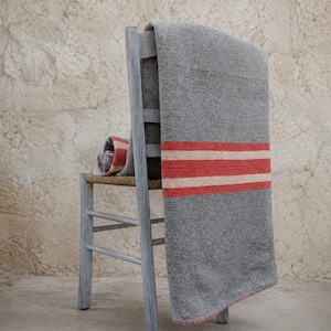 Può includere: Una sedia in legno grigio con seduta intrecciata presenta un tessuto grigio drappeggiato con due strisce orizzontali rosse e una striscia color crema tra di esse. Il tessuto pende dallo schienale, mostrando una superficie testurizzata e una parte ripiegata.