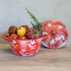 Peut inclure: Une marmite en métal émaillé rouge et blanc avec deux poignées latérales et un couvercle assorti. La marmite est décorée d'un motif floral blanc et contient plusieurs tomates dans des tons rouge, jaune et violet foncé, avec un brin de romarin vert.