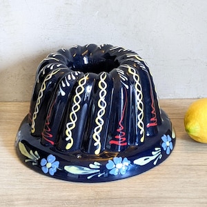 May include: A dark blue ceramic bundt cake pan featuring a fluted design with hand-painted yellow and white swirling lines, red accents, and a base decorated with light blue flowers and green leaves, positioned next to a single yellow lemon.