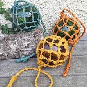 Op de afbeelding: Drie glazen visdrijvers in groen, oranje en geel, elk omhuld in een geknoopt net. De drijvers worden getoond op een verweerd houten oppervlak, met een getextureerde muur op de achtergrond. Elke drijver heeft een touw bevestigd.