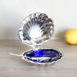 Peut inclure: Un plat en forme de coquille de palourde en argent avec un insert en verre bleu et une petite cuillère en argent. Le plat est ouvert et la cuillère repose sur le bord du plat.