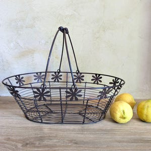 Puede incluir: Una cesta de alambre de metal marrón oscuro con asa y recortes de flores. La cesta de forma ovalada se encuentra sobre una superficie de madera, con tres limones amarillos a la derecha. La cesta está vacía.