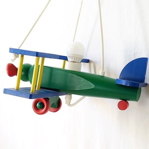 May include: A wooden airplane-shaped pendant light. The body is green, with blue, yellow, and red accents. The light bulb is white. The plane is suspended by a white cord.