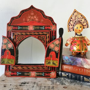 May include: A decorative, hand-painted, red and black mirror with floral and geometric designs. The mirror has two hinged doors. A wooden doll in traditional Russian attire stands on a stack of books next to the mirror.
