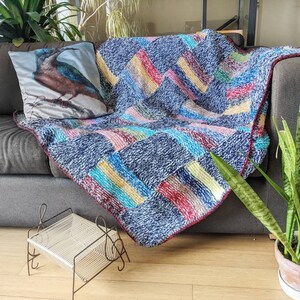 Op de afbeelding: Een blauwe en veelkleurige patchwork deken met een franje rand is over een grijze bank gegooid. Een kussen met een vogel illustratie is op de bank. Een plant in een pot staat op de vloer naast de bank.