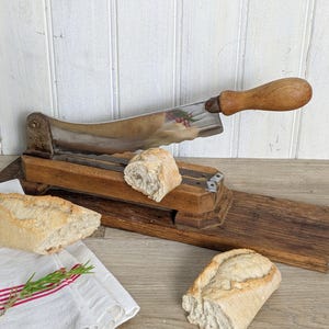 Peut inclure: Une trancheuse à pain en bois vintage dotée d'une longue lame métallique incurvée avec un manche en bois, fixée sur un socle en bois. Deux morceaux de baguette croustillante sont posés près de la trancheuse, avec un linge blanc à rayures rouges.