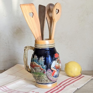 Peut inclure: Une chope à bière en céramique bleu foncé avec une anse, remplie d'ustensiles de cuisine en bois. La chope présente une scène peinte d'une femme et d'une maison. Un citron est posé sur un tissu à carreaux rouge et blanc.
