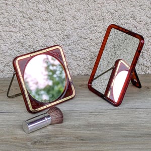 Op de afbeelding: Twee make-upspiegels met schildpadpatroon frames en een make-upkwast. De ene spiegel is vierkant met een cirkelvormig glazen midden en goudkleurige rand, terwijl de andere rechthoekig is. Beide spiegels hebben metalen standaards voor rechtop staan.
