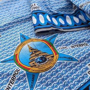 Peut inclure: Un tissu à motifs bleus et blancs présentant un emblème en forme d'étoile avec le texte 71st GENERAL ASSEMBLY ICPO-INTERPOL YAOUNDE, 21-24 Oct. 2002. Le motif inclut les mots ASIE, AFRIQUE, EUROPE, OCEANIE et AMERIQUE autour de l'étoile.