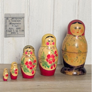 May include: A set of four wooden nesting dolls with painted faces, red headscarves, and floral patterns on their bodies. A small rectangular paper label is attached to the wall, featuring Russian text including the year 1965 and the word Matreshka.