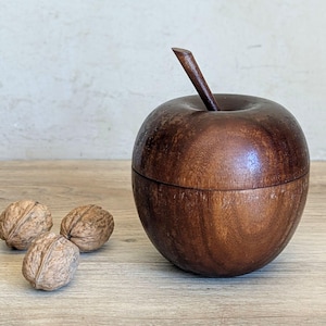 May include: A dark brown wooden apple-shaped container with a stem. The container is on a wooden surface, with three walnuts. The container is likely a decorative storage item, with a lid that fits snugly.