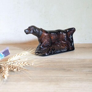 May include: A brown glass dog figurine with detailed fur texture. The dog is in a resting pose, with its head and tail slightly raised. The figurine is on a wooden surface, with wheat stalks and a small piece of paper in the background.