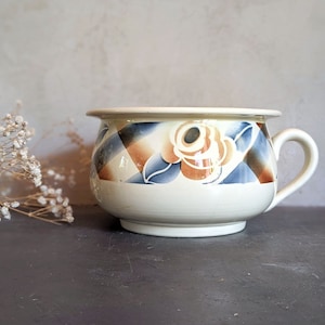 Puede incluir: Un orinal de cerámica de color crema con un borde ancho, una sola asa lateral y una banda decorativa alrededor del centro. La banda muestra un patrón repetitivo de formas de diamante en azul y marrón intercaladas con motivos florales.