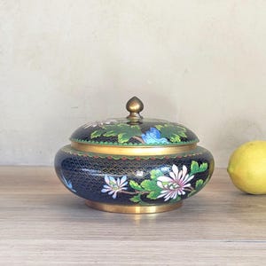 May include: A round, black cloisonné lidded jar featuring a detailed floral pattern with white, pink, and blue petals and green leaves. The jar has a brass rim, a brass base, and a small, pointed brass finial on the center of the lid.