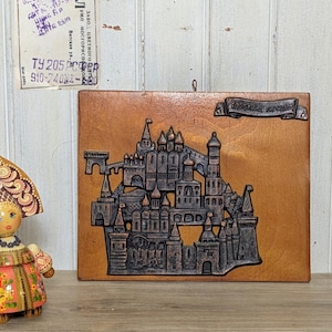 May include: A rectangular wooden wall plaque featuring a dark brown relief sculpture of a fortress with multiple towers and domes. A small banner at the top right contains Cyrillic text. A small portion of a painted wooden doll is visible on the left.