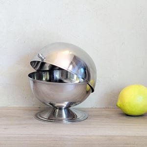 May include: A spherical stainless steel serving bowl with a polished finish, featuring a hinged lid that opens to reveal the interior. The bowl is supported by a circular pedestal base and is positioned next to a single yellow lemon.