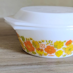 May include: White, round casserole dish with a lid. The dish features a floral pattern in orange, yellow, and green. The word "codec" is printed on the side of the dish.