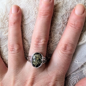 Puede incluir: Un anillo de plata con un engaste de resina negra de forma ovalada que presenta un diseño de flor blanca prensada.