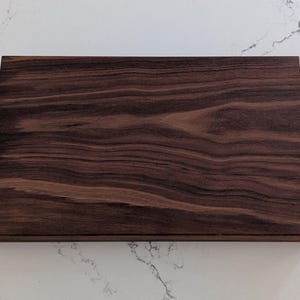May include: A rectangular wooden cutting board with a rich, dark brown color and visible wood grain. The board has a smooth surface and a beveled edge. The cutting board is made of hardwood.