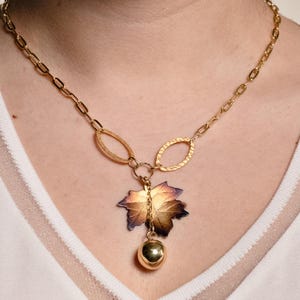 May include: A gold-toned chain necklace featuring two textured oval links connected to a central pendant. The pendant consists of a multi-colored maple leaf in shades of bronze, purple, and gold, with a small, smooth, polished gold sphere hanging below.