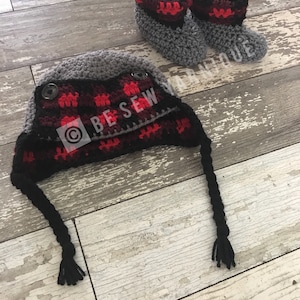 May include: A handmade crochet hat and matching booties set. The hat is gray with a red and black plaid design on the front, secured with two buttons. The booties are gray with red and black trim. The set is designed for babies.