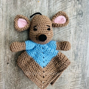May include: A brown crocheted mouse with blue and brown blanket. The mouse has pink ears and black eyes.