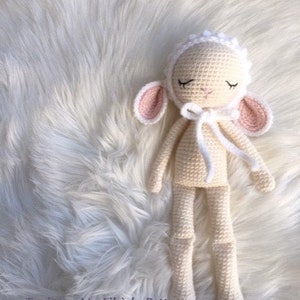 Puede incluir: Una muñeca de cordero blanco de crochet con los ojos cerrados, que lleva una boina blanca de crochet. La muñeca está tumbada sobre una superficie blanca y esponjosa. El texto "Leonie la brebis" está escrito encima de la muñeca y "Toujours Un Fil à la Patte" está escrito debajo de la muñeca.