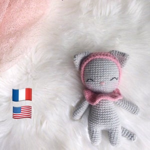 May include: A gray crocheted cat toy with pink ears, a pink hat, and a pink ruffle collar. The cat is lying on a white fluffy surface. The text "Collette le Capu' Choux" is at the top of the image and the text "Toujours Un Fil à la Patte" is at the bottom of the image.