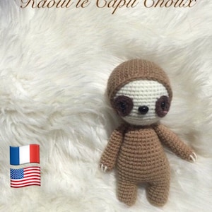 May include: A brown crocheted sloth toy with a white hood. The toy is sitting on a white fluffy surface. The text "Raoul le Capu' Choux" is at the top of the image. The text "Toujours Un Fil à la Patte" is at the bottom of the image. The flags of France and the United States are in the lower left corner of the image.