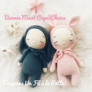 Puede incluir: Dos muñecas de conejo de crochet, una en rosa y otra en azul turquesa, tumbadas sobre una superficie blanca y esponjosa. Las muñecas tienen ojos y narices bordados y llevan atuendos a juego. El texto "Bunny Maxi Capu' Choux" está escrito encima de las muñecas, y el texto "Toujours Un Fil à la Patte" está escrito debajo de las muñecas.
