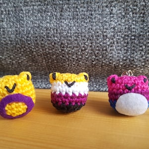 May include: Three crocheted frog keychains in yellow, non-binary, and bisexual pride flag colours. The frogs have black stitched eyes and smiles. The yellow frog has a purple mouth. The non-binary frog has a white, purple, and black design. The bisexual frog has a pink, blue, and white design.