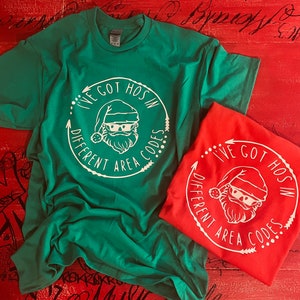 May include: Two t-shirts, one green and one red, with a white graphic of Santa Claus wearing a hat and the text "I'VE GOT HOS IN DIFFERENT AREA CODES" in a circular design. The shirts are on a red wooden surface.
