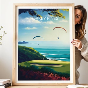 Puede incluir: Un póster enmarcado con una ilustración colorida de un campo de golf con vistas al océano. El póster incluye el texto "TORREY PINES, California". Tres parapentes vuelan en el cielo sobre el océano.