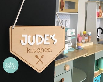 May include: A wooden sign with the text "JUDE'S kitchen" in white letters with a brown outline. The sign is hanging from a white rope and has a brown wooden spoon and fork crossed underneath the text. The sign is hanging in front of a black chalkboard.